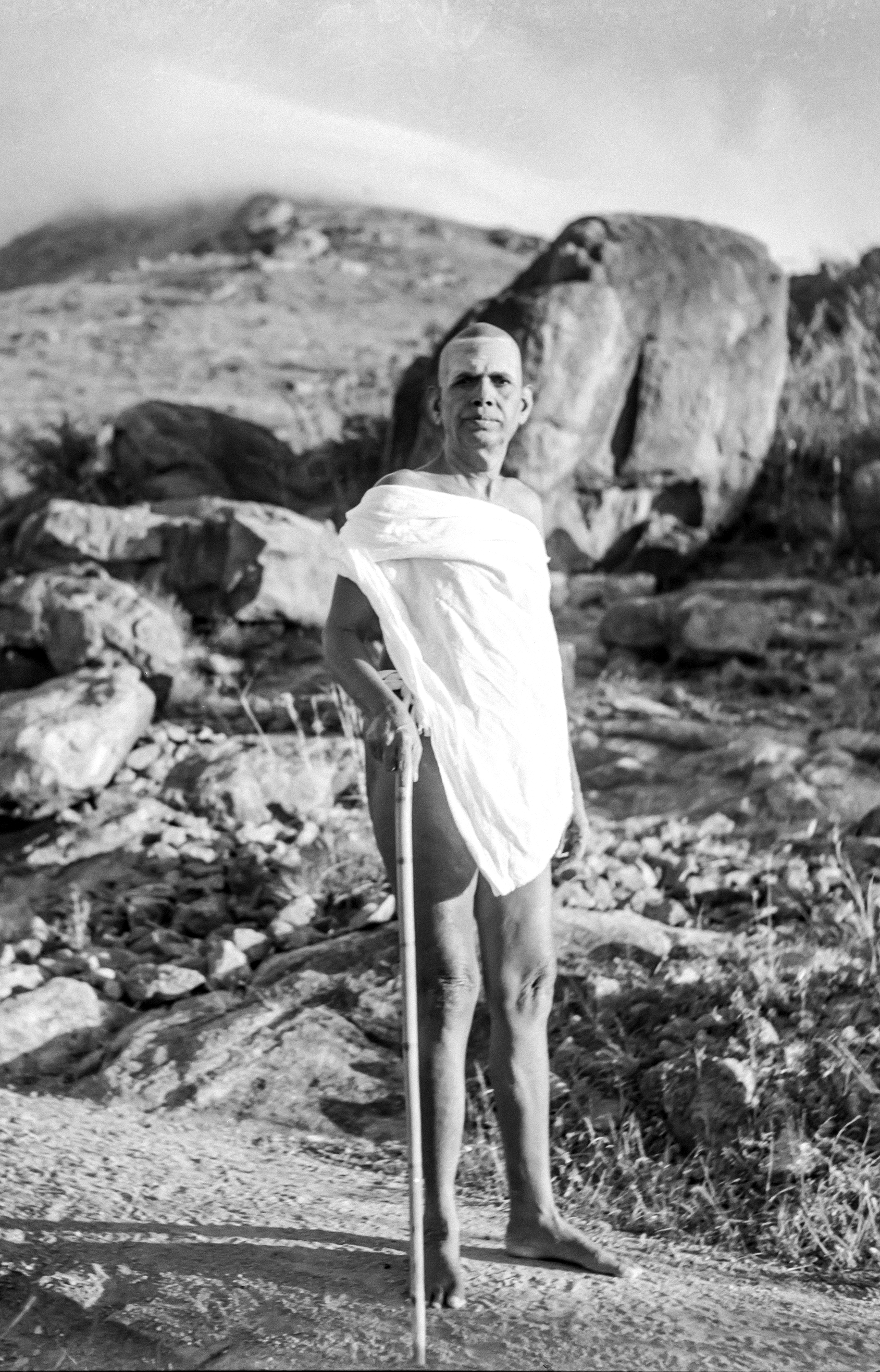 Bhagavan Sri Ramana Maharshi standing with a walking staff on the rocky slopes of Arunachala