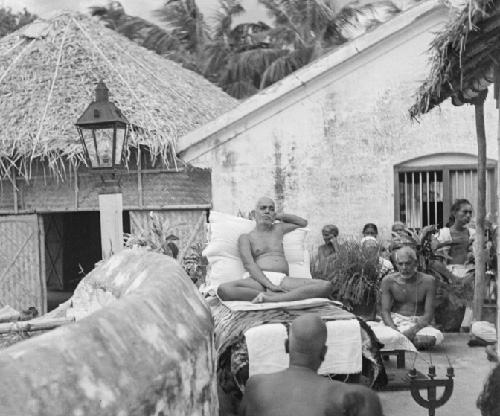 Bhagavan Sri Ramana Maharshi seated on a couch outside the thatched-roof hall at Sri Ramanasramam surrounded by devotees sitting in silence