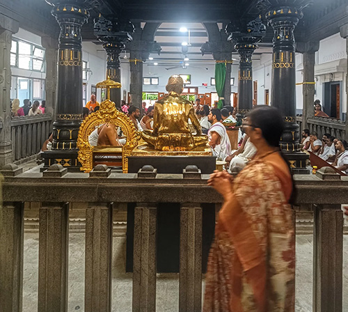 A devotee in an orange sari performing pradakshina around the samadhi shrine of Bhagavan Ramana, with polished black stone pillars and the gold-plated statue of Sri Bhagavan